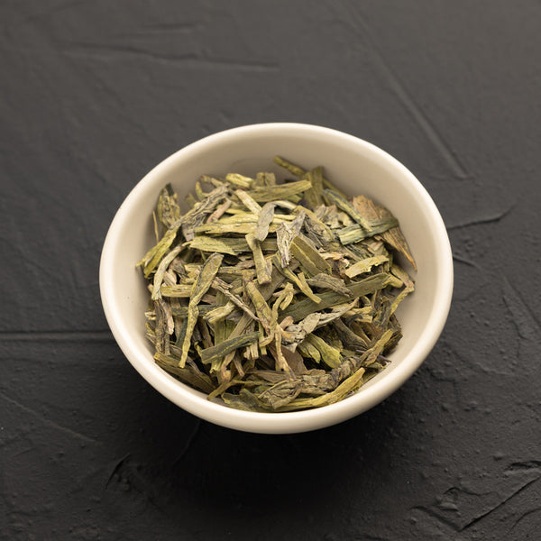 White bowl filled with green Long Jing tea leaves on a dark gray background
